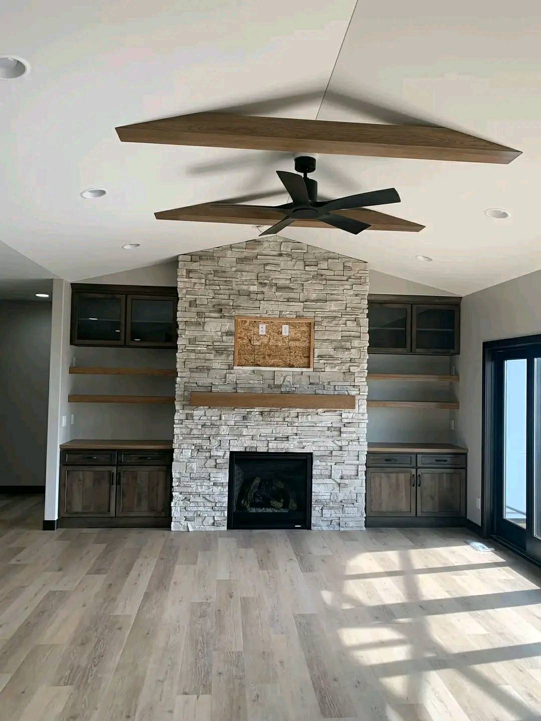 Modern living room with a stone fireplace and wooden mantel, flanked by built-in shelves and cabinets. A ceiling fan and large windows add to the airy feel.
