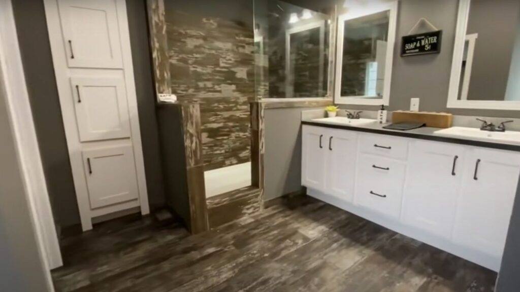 Modern bathroom with rustic elements, featuring a glass shower with wood-patterned tiles, dual white vanities, gray walls, and dark wood flooring.