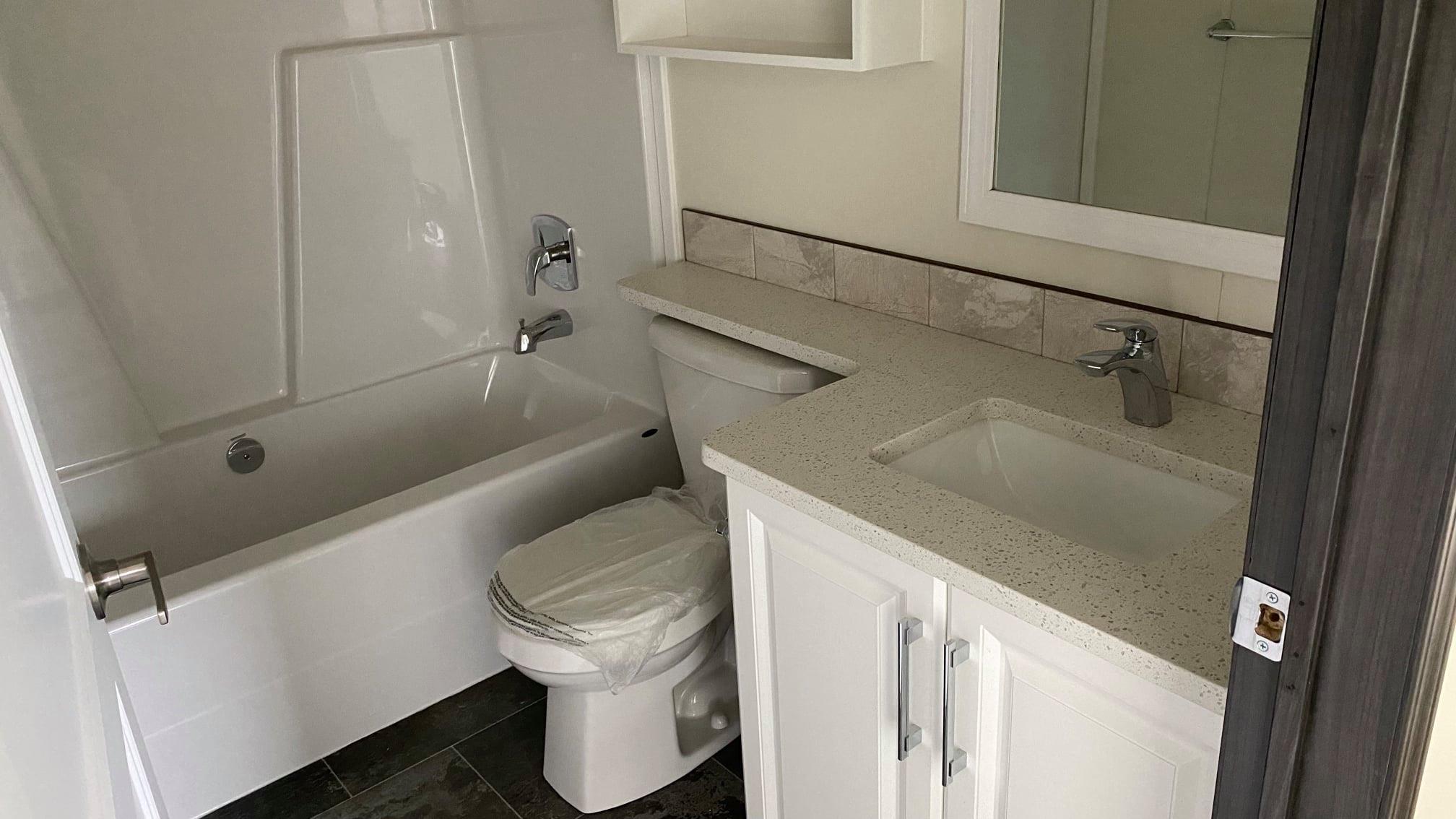 Compact modern modular home bathroom interior under construction featuring white acrylic bathtub with tiled surround, white toilet with protective covering, beige speckled quartz vanity countertop, undermount rectangular sink, brushed nickel faucet and cabinet pulls, white shaker-style vanity cabinets, large wall mirror, open shelving above toilet, neutral beige walls, dark gray floor tiles, bright clean contemporary manufactured home ensuite bathroom view