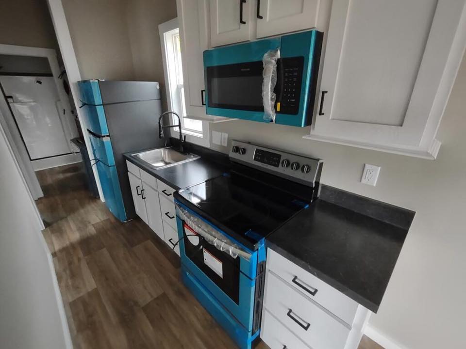 Modern kitchen with light wood flooring, featuring a blue-covered fridge and oven. A black countertop, white cabinets, and a stainless steel sink enhance the contemporary look.