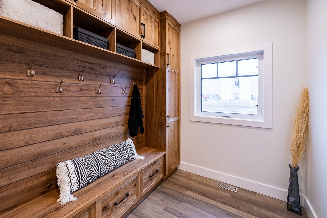 Rustic mudroom with wooden storage benches, hooks, and upper cabinets. A patterned cushion sits on the bench. Bright window, pale walls. Cozy feel.
