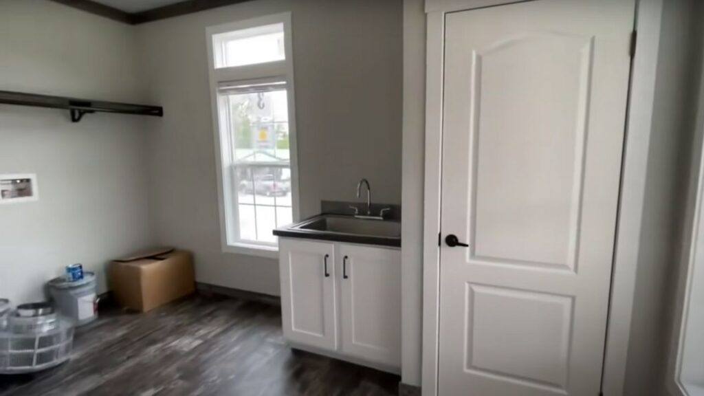A small, modern utility room with a sleek white cabinet and sink, dark wood flooring, a high window, and a door. The tone is clean and minimalistic.