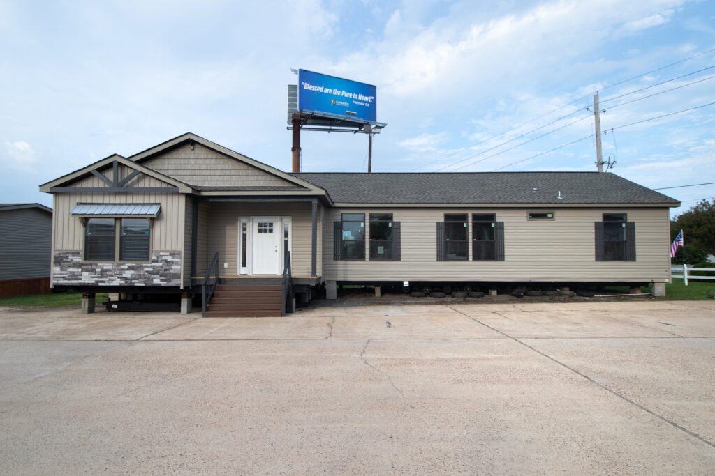 Single-story modular home on a paved lot with a porch entrance, gray siding, and stone accents. A billboard above reads "Blessed are the Pure in Heart."