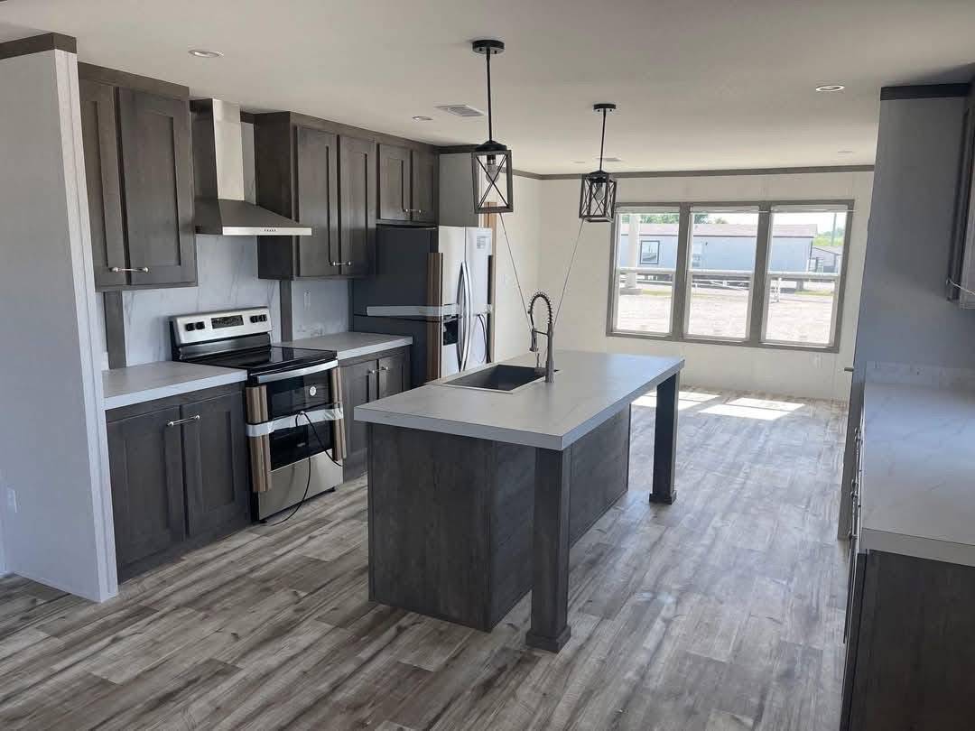 Modern open-concept kitchen with spacious grey quartz island, dark grey shaker cabinets, stainless steel Frigidaire appliances, black farmhouse pendant lights, large panoramic windows, luxury vinyl plank flooring, and bright natural daylight – perfect new construction modular home kitchen interior 2025