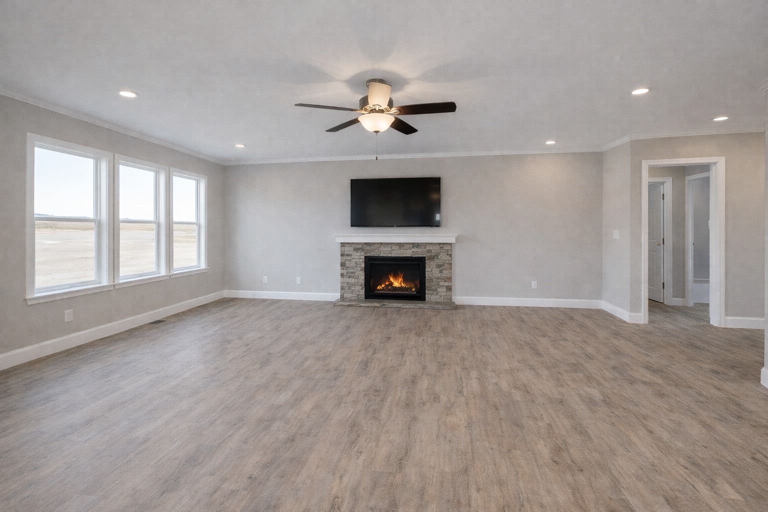 Spacious living room with wood floors, gray walls, and large windows. A stone fireplace with a TV above and a ceiling fan enhance the cozy atmosphere.