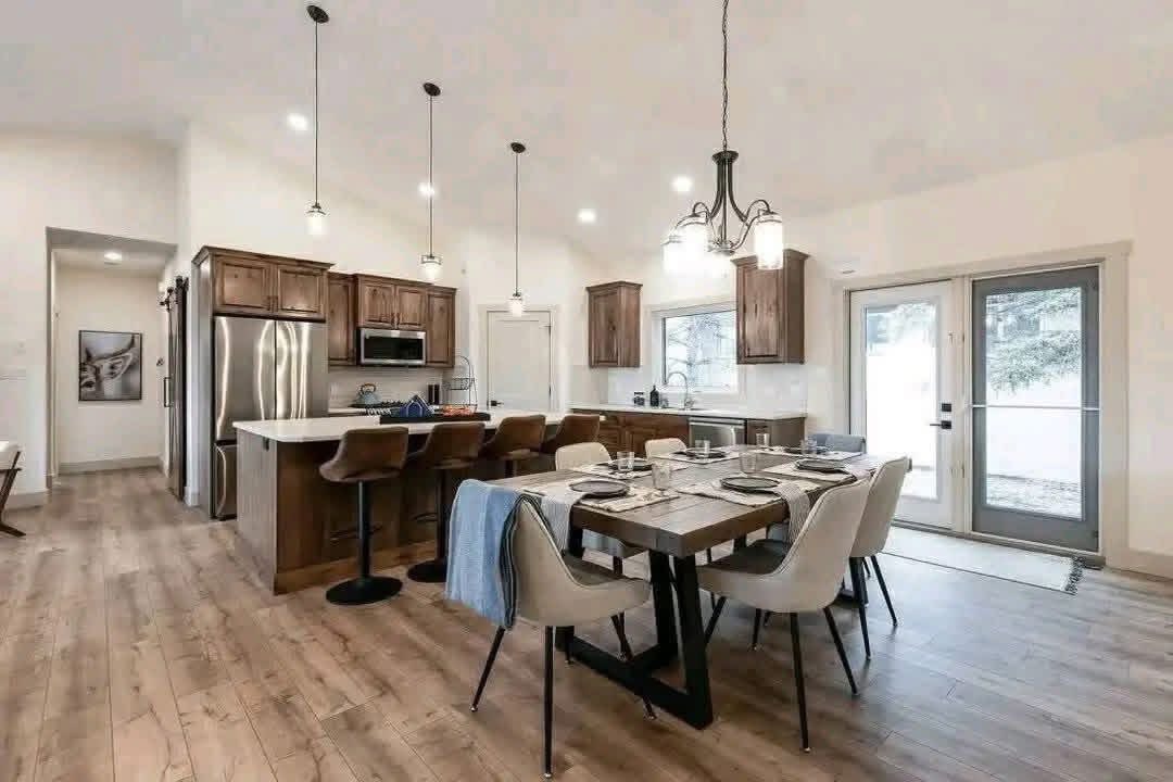 Modern kitchen and dining room with a cozy feel. Features wooden floors, a large table set for six, a kitchen island with barstools, and pendant lighting.