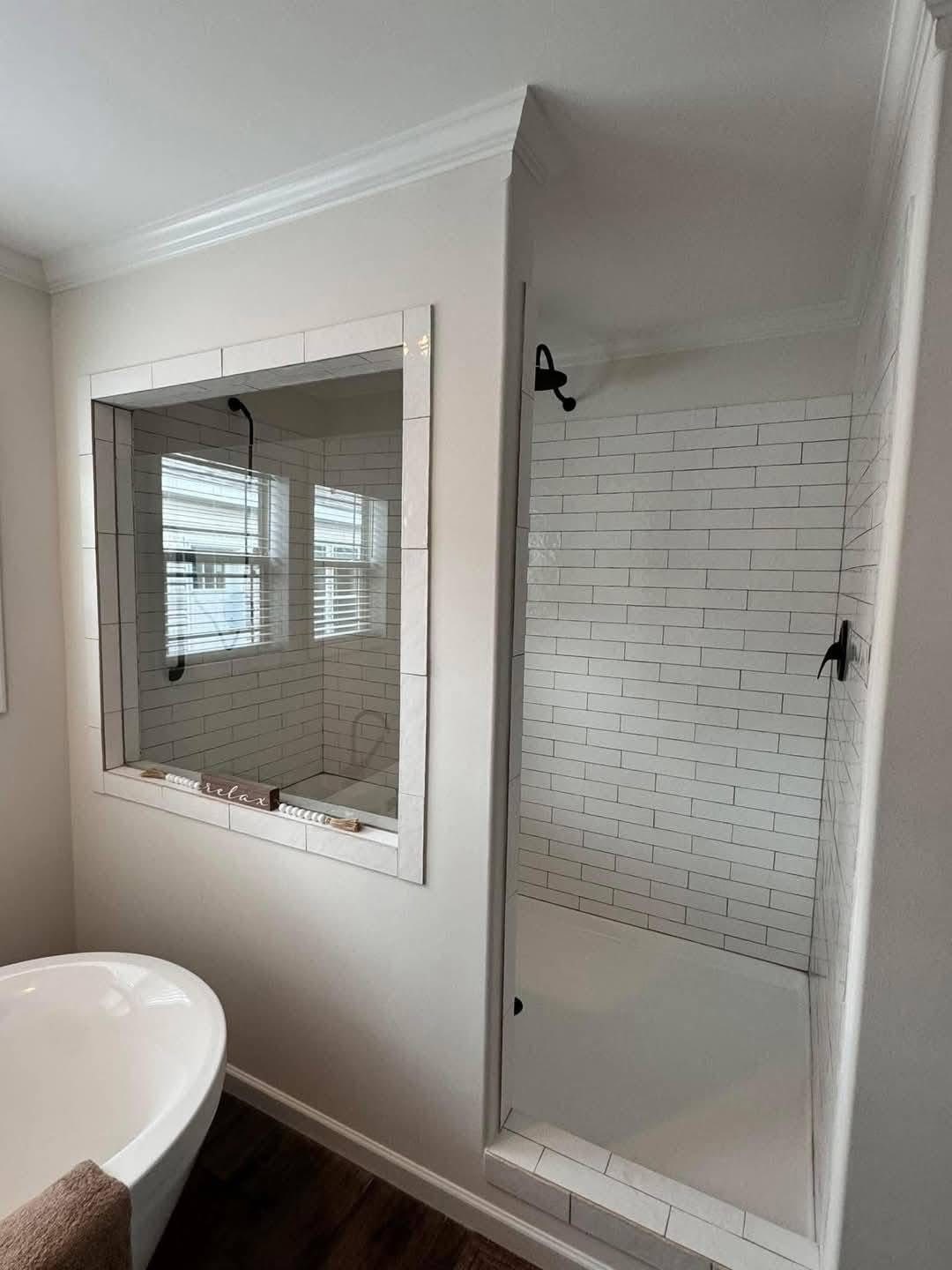 A bright bathroom with a white bathtub, dark wood floor, and a glass-enclosed shower featuring white subway tiles. The atmosphere is clean and serene.