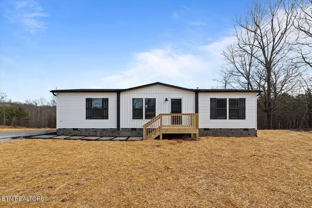 Modern charming single-story modular ranch home exterior featuring crisp white board-and-batten siding with bold black trim accents, black-framed double-hung windows with matching black shutters, sleek black front door under simple gable porch, wooden handrail staircase with natural cedar steps and landing, dark metal roof edges, raised foundation on concrete block piers with black skirting, spacious manufactured bungalow facade set on open grassy lot against clear blue sky with scattered clouds and bare winter trees in background