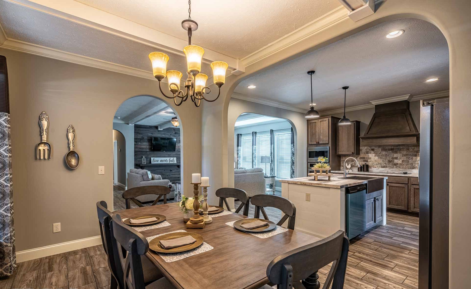Warm and inviting dining room with a wooden table set for four. A chandelier hangs above, leading into a cozy kitchen with dark cabinets and modern appliances.