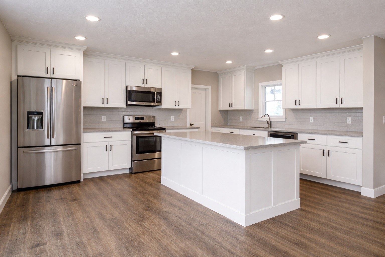 Modern kitchen with stainless steel appliances, white shaker cabinets, and a large central island. Wood flooring adds warmth to the bright space.