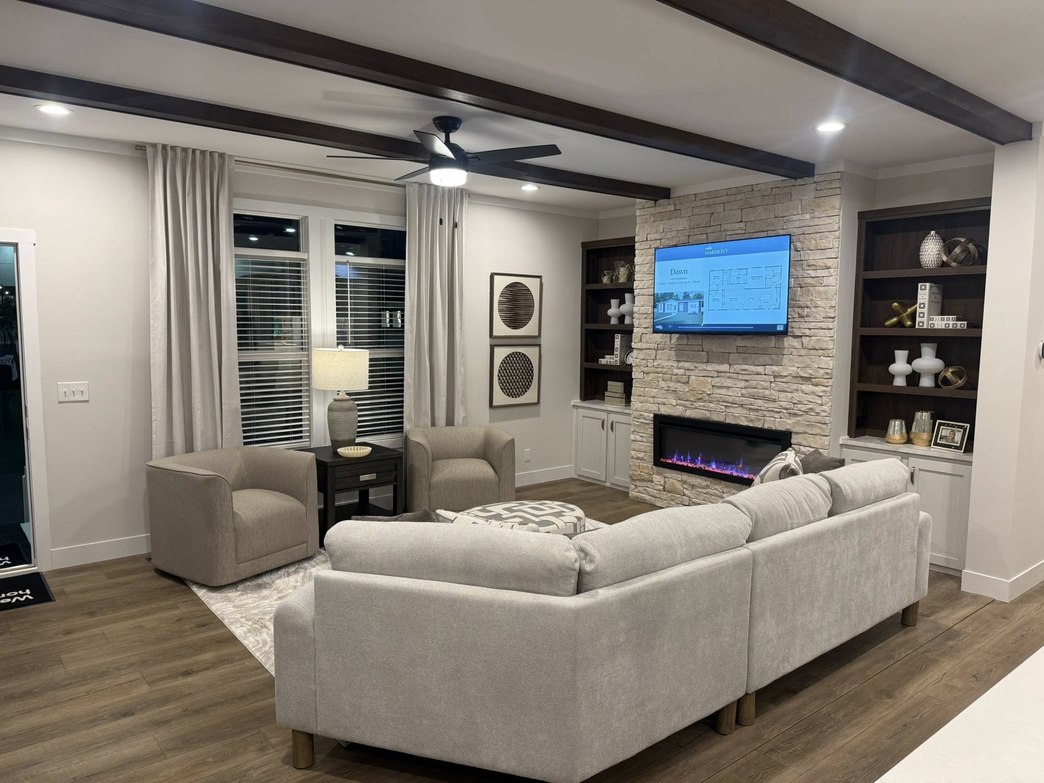 Modern living room with a gray L-shaped sofa, stone fireplace, wall-mounted TV, and wooden shelves. Soft lighting creates a cozy atmosphere.