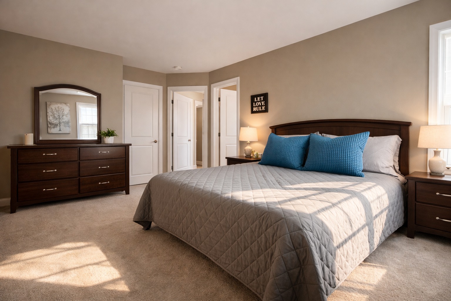 A cozy bedroom with a beige color scheme features a large bed with a gray quilt and blue pillows. A wooden dresser with a mirror is on the left, and a nightstand with a lamp is on the right. Sunlight streams through a window, creating a warm and inviting atmosphere.