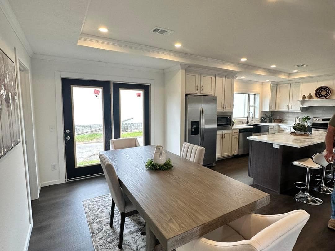 Modern kitchen and dining area with a wooden table, white upholstered chairs, and French doors. Stainless steel appliances and dark wood flooring.