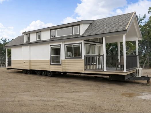 Charming white and tan tiny house on wheels with a covered porch, multiple windows, and a pitched roof, parked on a gravel lot under a partly cloudy sky, showcasing modern mobile living solutions.