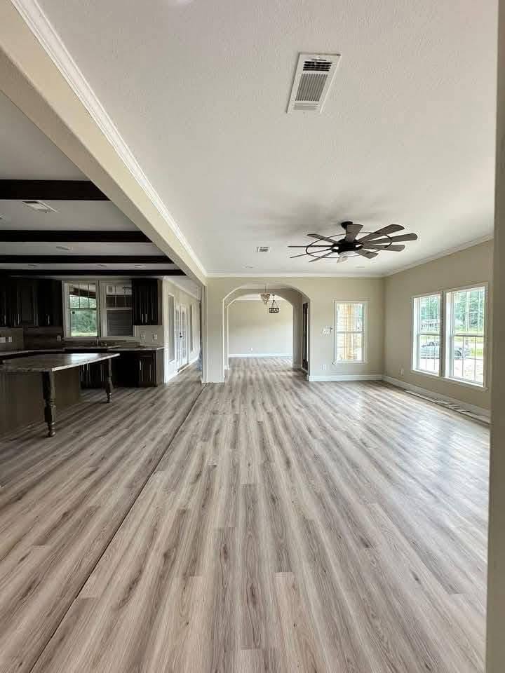 Spacious open-plan living room with light wood flooring, large windows, and a ceiling fan. Adjacent kitchen has dark cabinets, creating a modern feel.