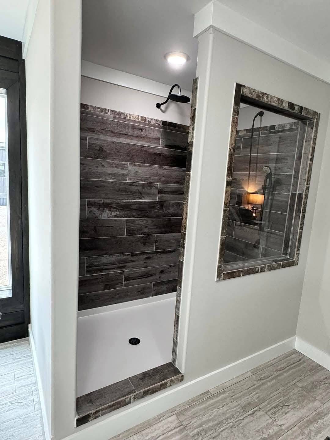 Modern bathroom with marble-accented open shower, dark wood tiles, and a glass panel reflecting light. Sleek, minimalist design with a calming ambiance.