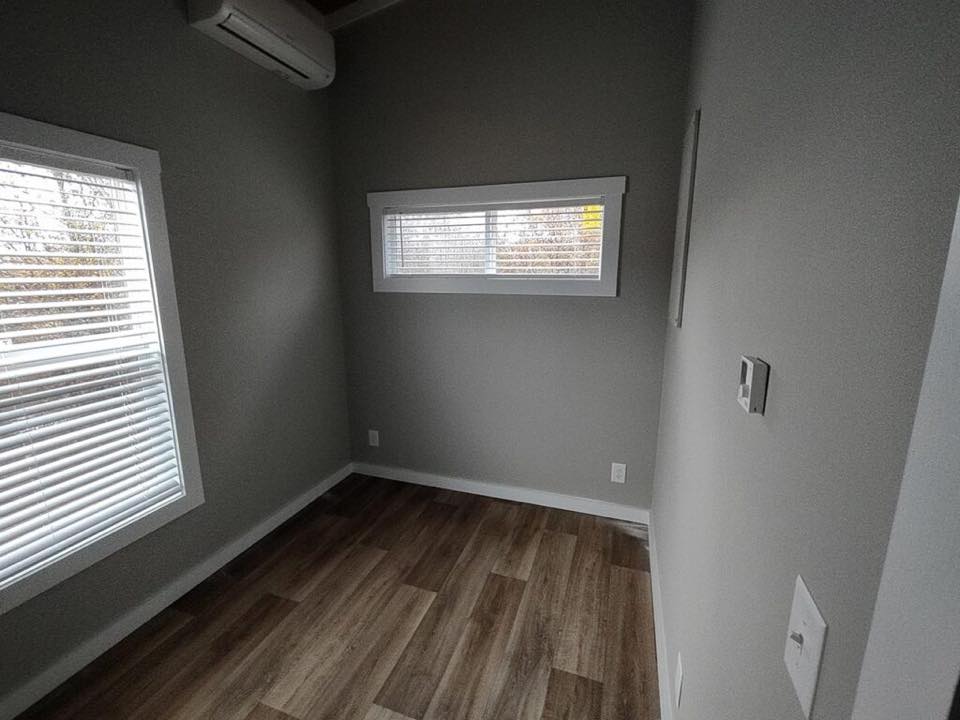 Small room with light gray walls and wood-patterned floor. Two windows with blinds bring in soft natural light. An air conditioner is mounted on the wall.