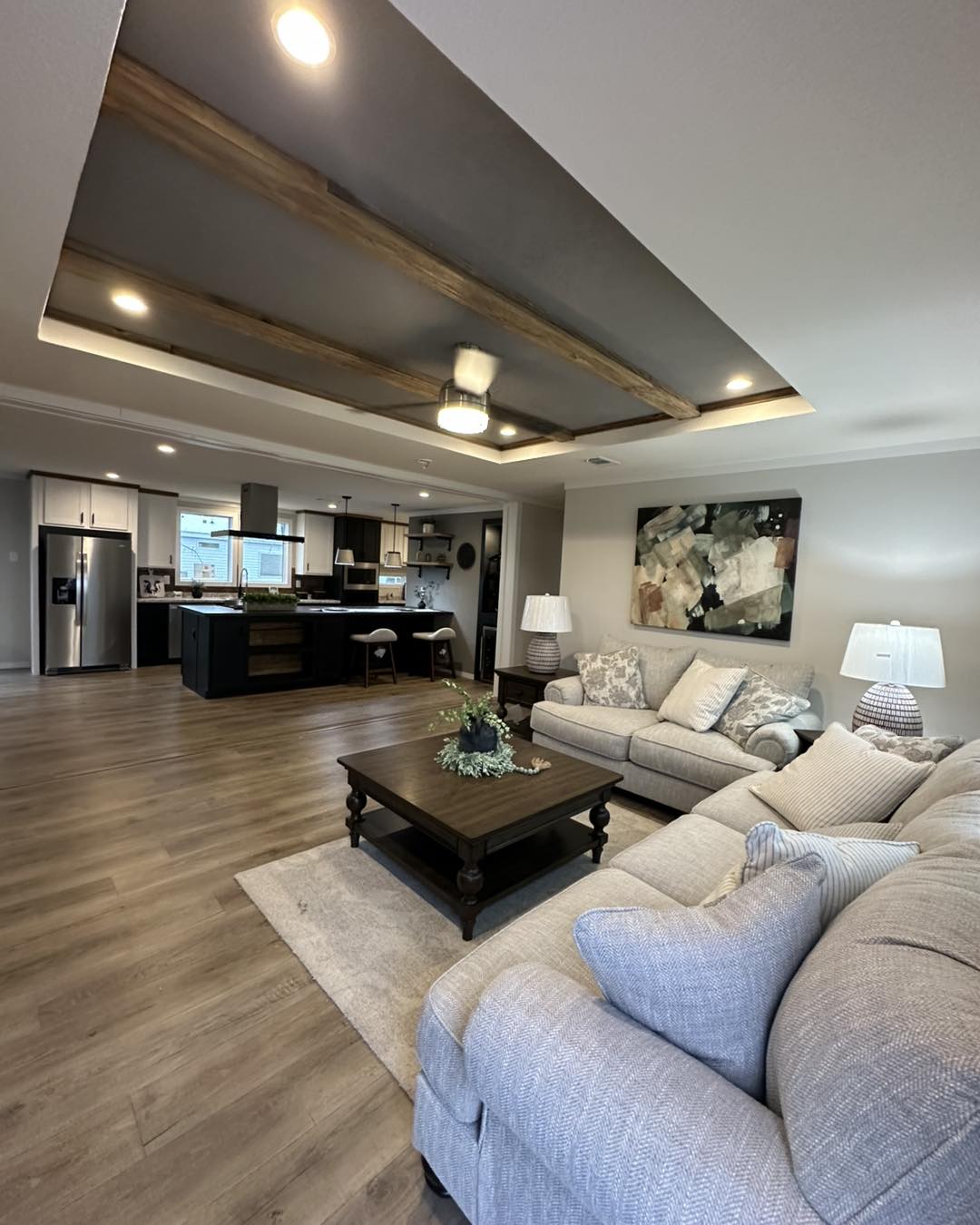 A modern, open-plan living room and kitchen with a gray sectional sofa, accent pillows, abstract wall art, and a wooden coffee table on a rug. Warm lighting and a cozy ambiance.