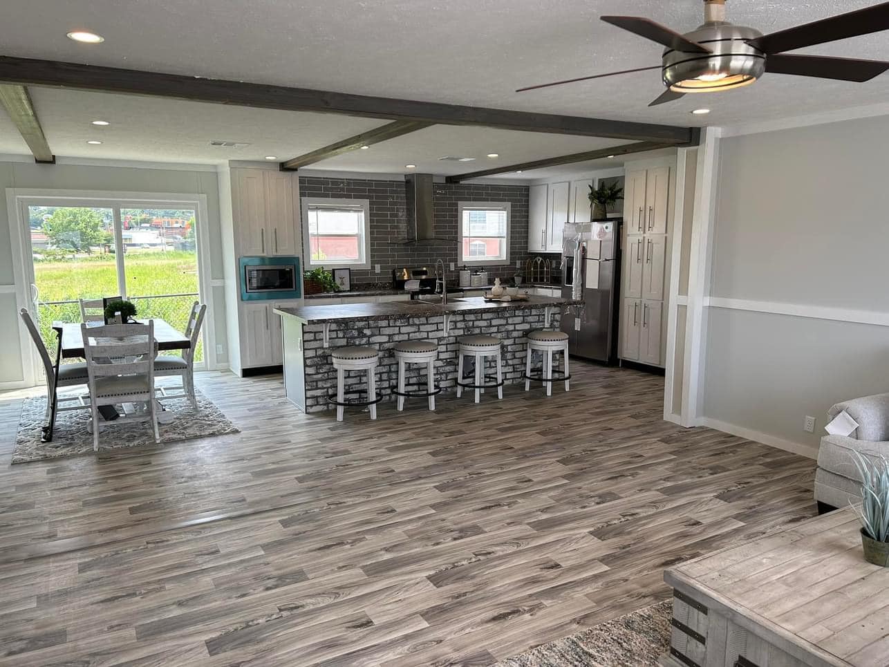 Modern open-concept kitchen and dining area with light wood flooring, a kitchen island with four stools, stainless steel appliances, and a dining table by large windows.