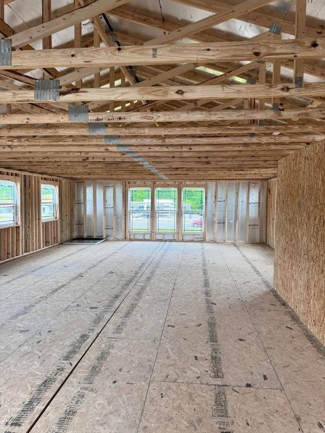 Open-concept unfinished room with wood framing and exposed beams. Large windows line the walls, allowing natural light to brighten the space.