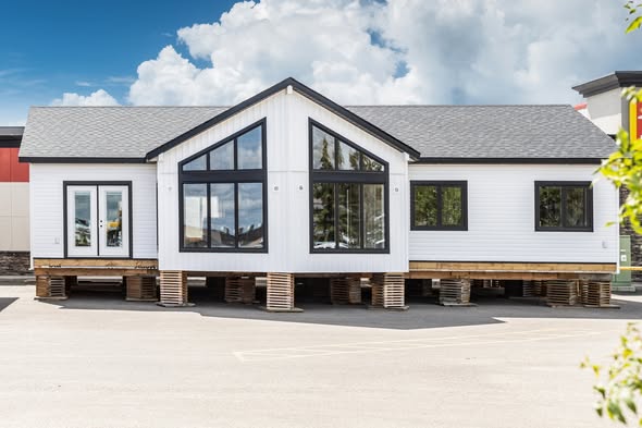 White modular home on wooden blocks, with large windows and a gray roof, set against a backdrop of cloudy sky. The scene is calm and orderly.