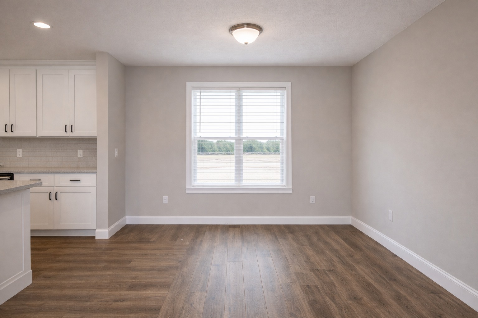 Empty room with wooden flooring, beige walls, and a central window with blinds. Left side features white kitchen cabinets and a counter. Bright, neutral ambiance.