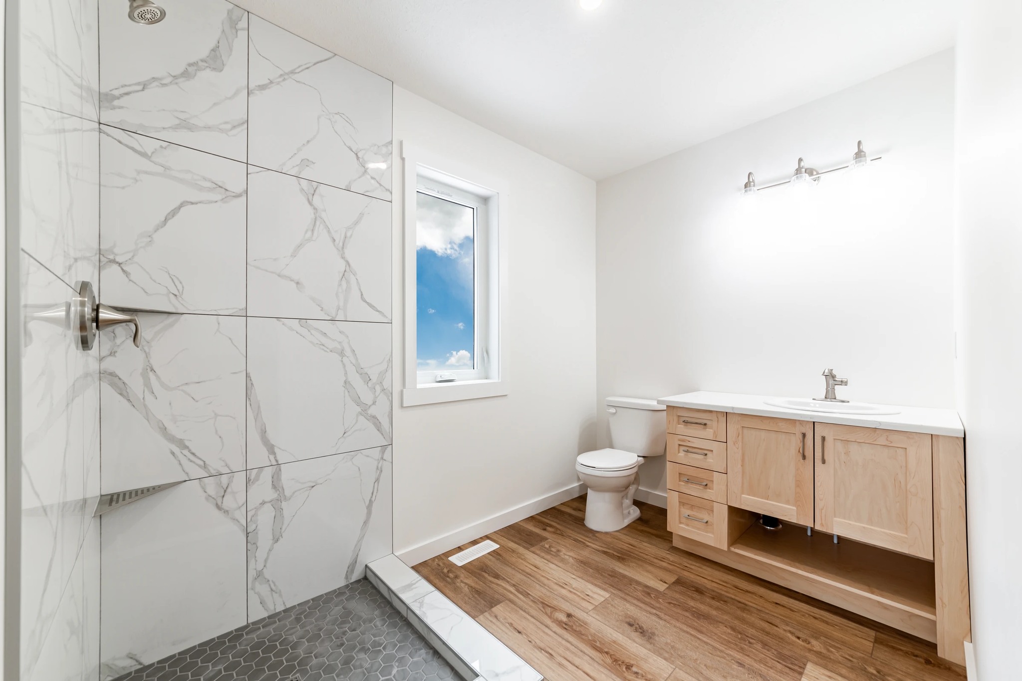A modern bathroom with a white marble-tiled shower, a wooden vanity, and a toilet. The window shows a blue sky, creating a bright, clean atmosphere.