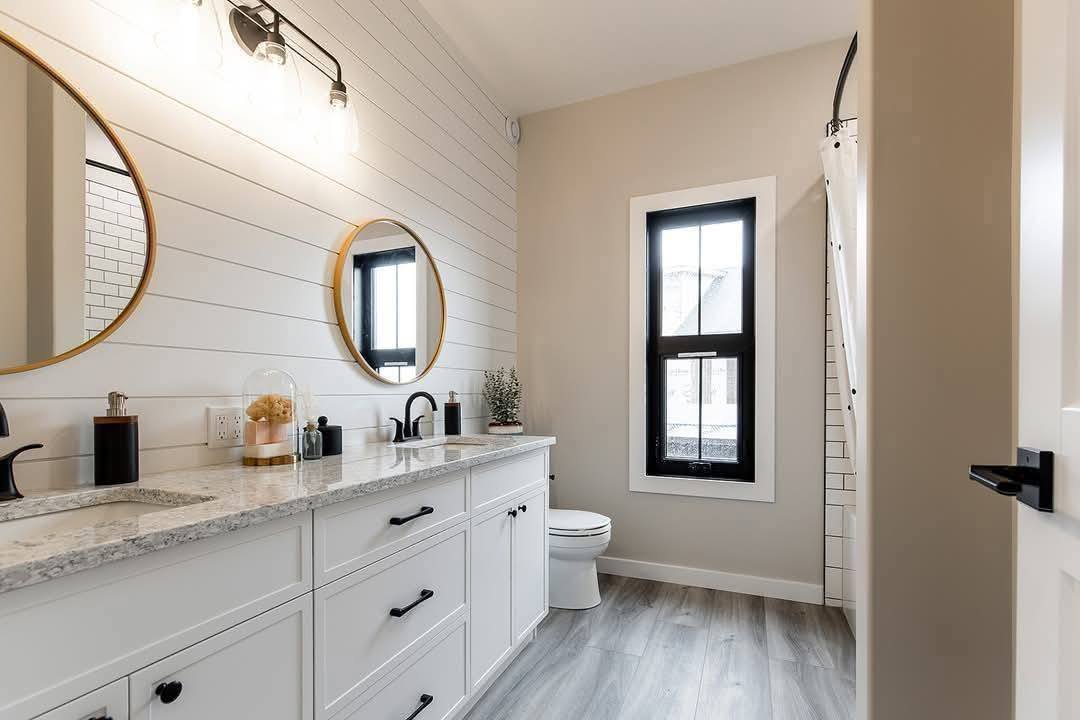 Modern bathroom with light wood flooring, white vanity cabinets, and round mirrors. Black fixtures complement the neutral tones. Bright and serene ambiance.