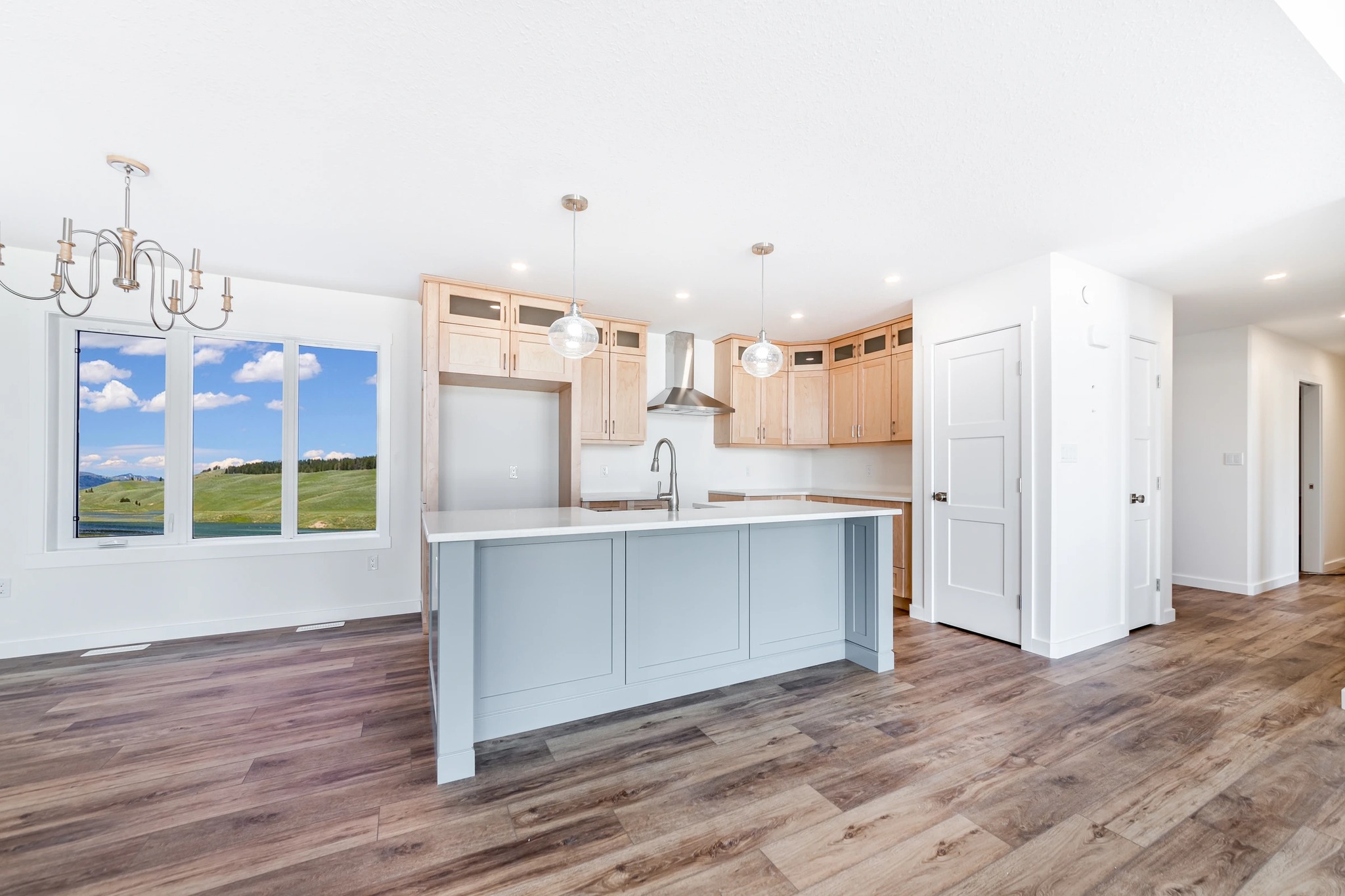 Spacious, bright kitchen interior with light wood cabinets, large blue island, pendant lights, and wood flooring. Window shows clear sky and fields.