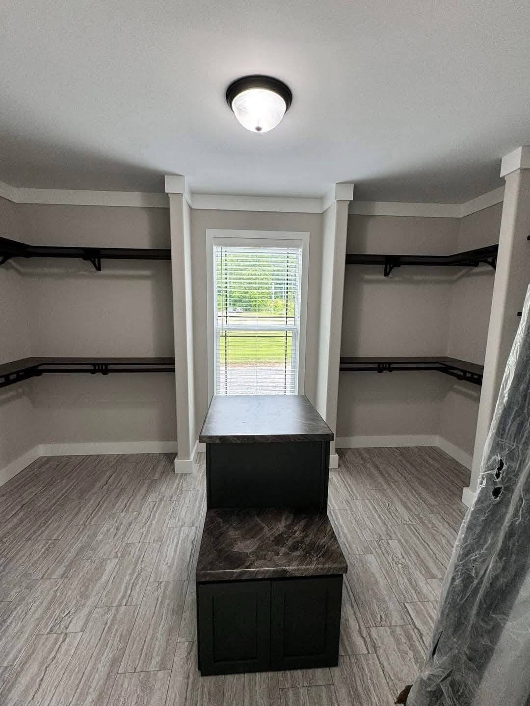 Spacious walk-in closet with gray flooring, dark shelves, and a window with blinds. Central dark wood bench. Bright, minimalistic design.