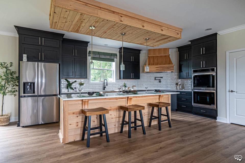 Modern kitchen with dark cabinets, stainless steel appliances, and a wooden island with three stools. Light wood floors and pendant lights create a cozy, stylish ambiance.