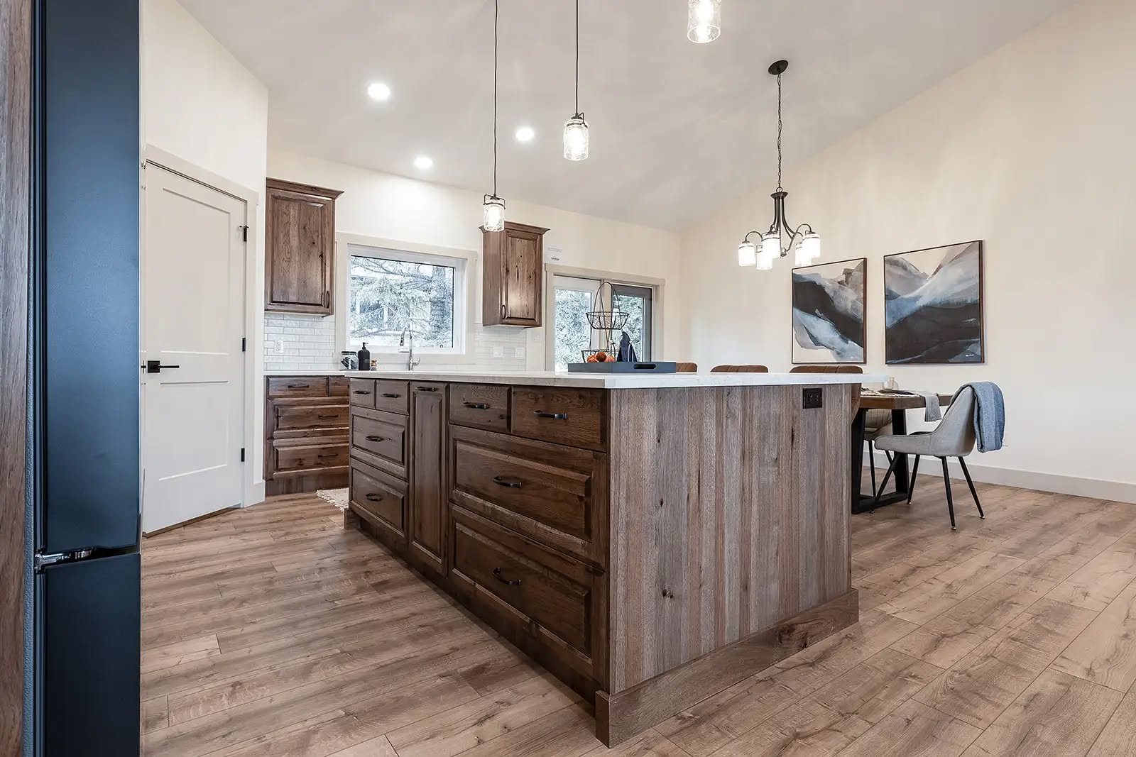 A spacious kitchen with a rustic wood island and modern lighting. It features hardwood floors, white walls, and a dining area with abstract art, creating a warm, inviting ambiance.