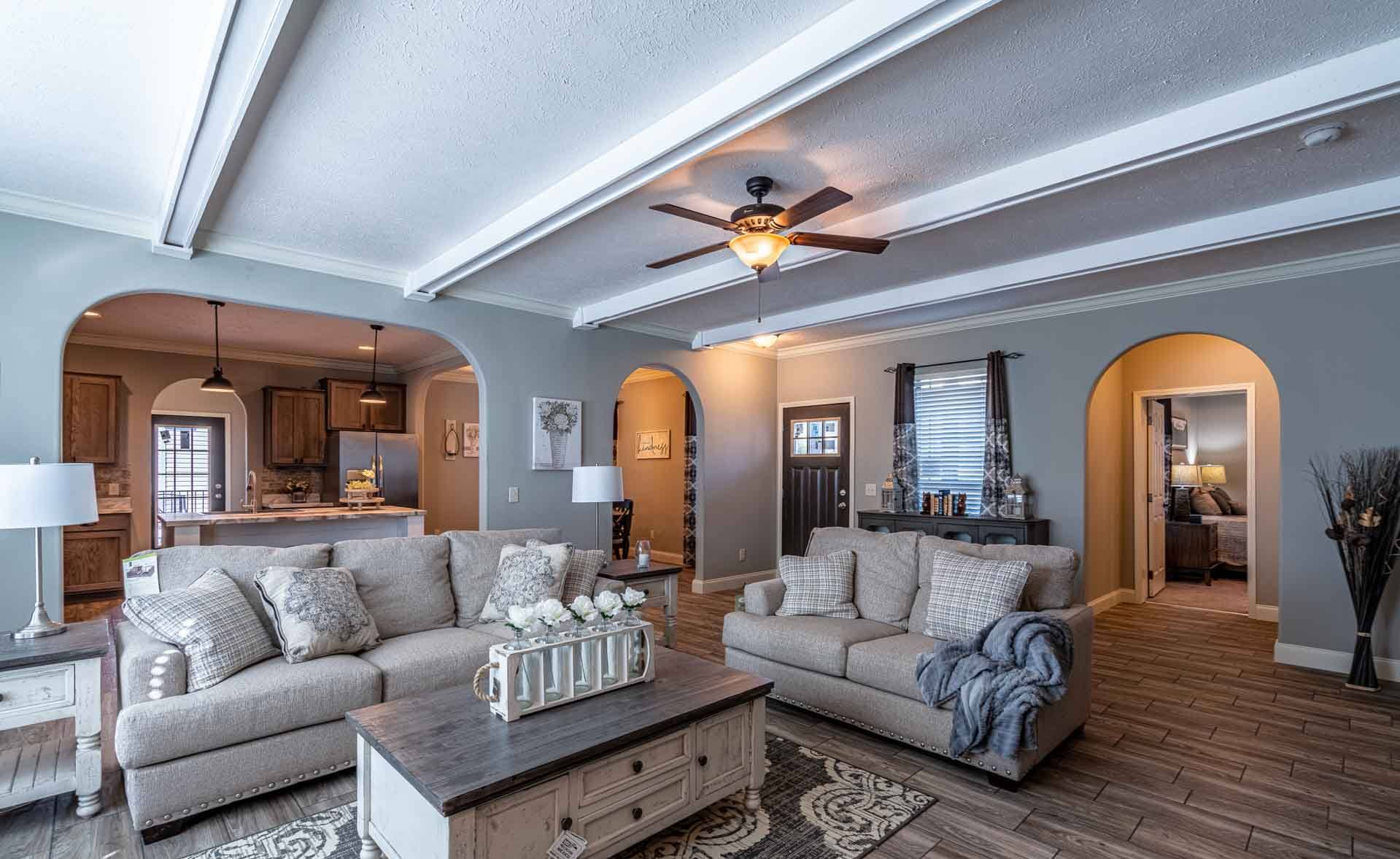 Spacious living room with beige sofas, wooden coffee table, and white decor accents. Arched doorways lead to a kitchen and hallway. Warm, cozy ambiance.
