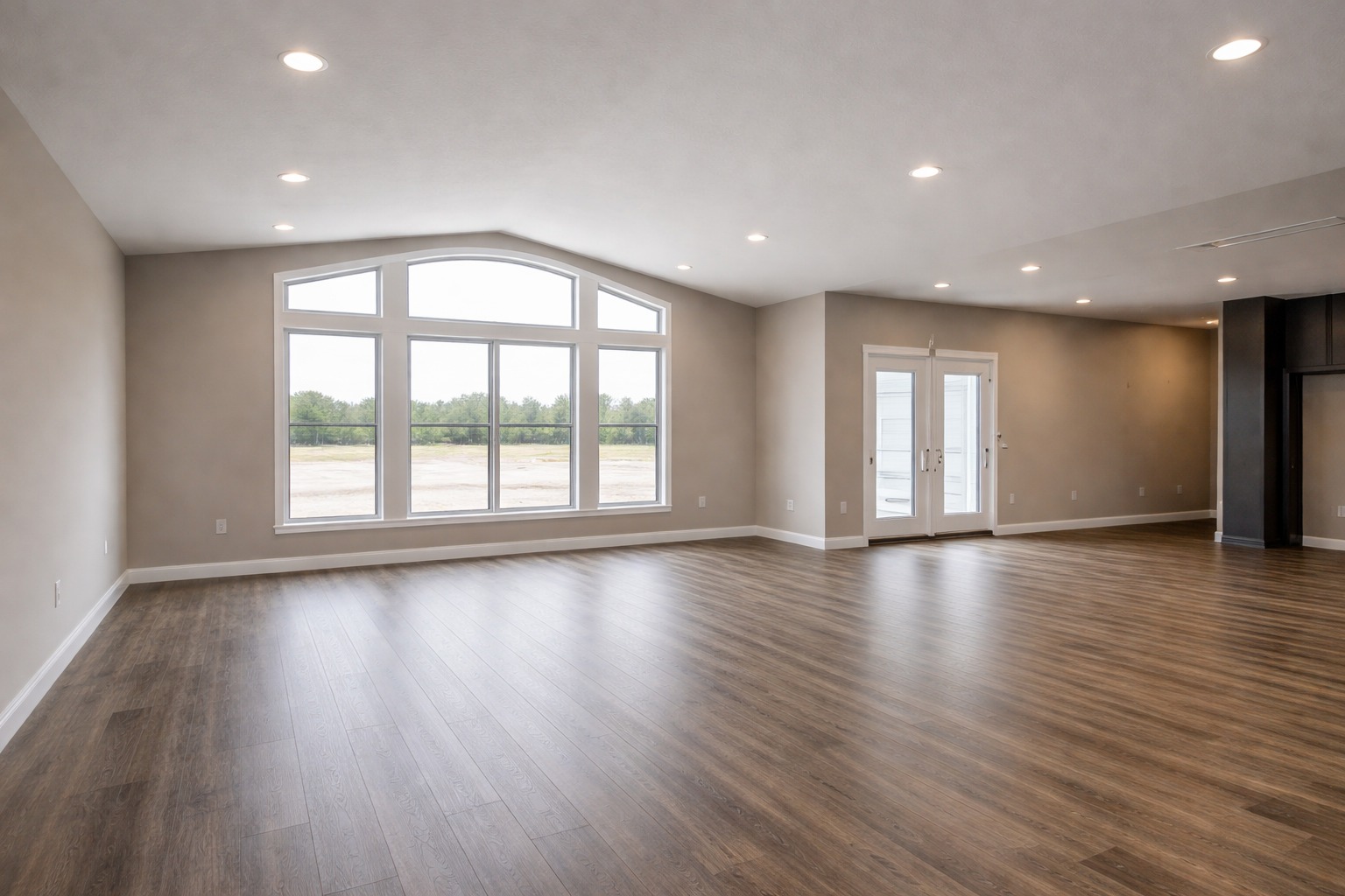 Spacious, empty room with light wood flooring, large arched windows showing greenery, beige walls, recessed lighting, and glass double doors. Calm ambiance.