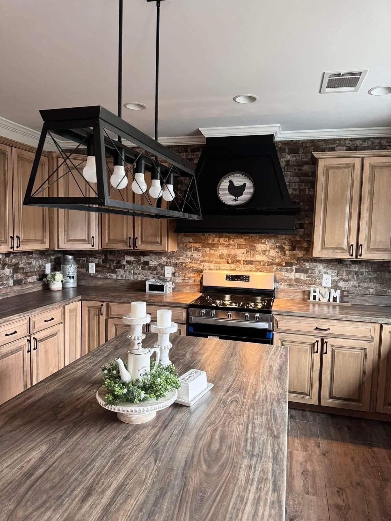 Rustic kitchen with wooden cabinets and a stone backsplash. A large island features candles and a plant. Black pendant lights add a cozy, farmhouse feel.