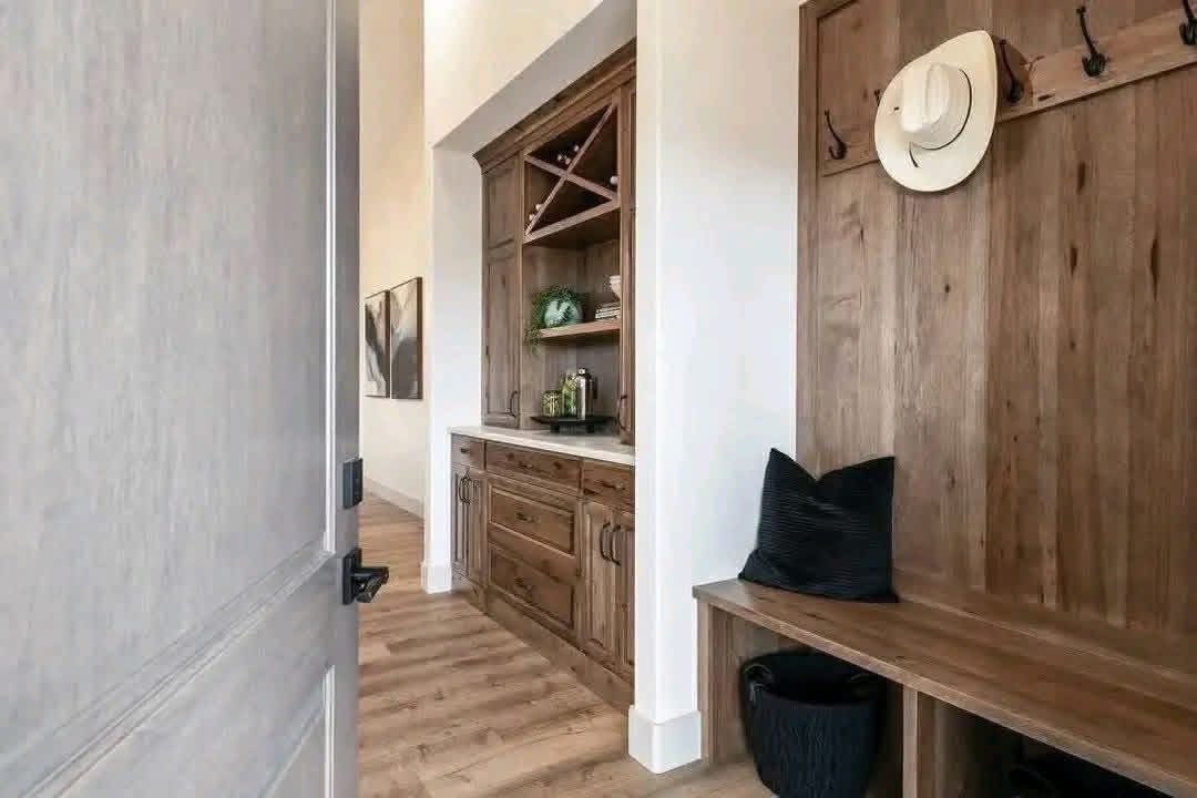 A rustic hallway features wood paneling and a bench with black cushion. A hat hangs on a hook. Wooden cabinetry with shelves holds decor items.
