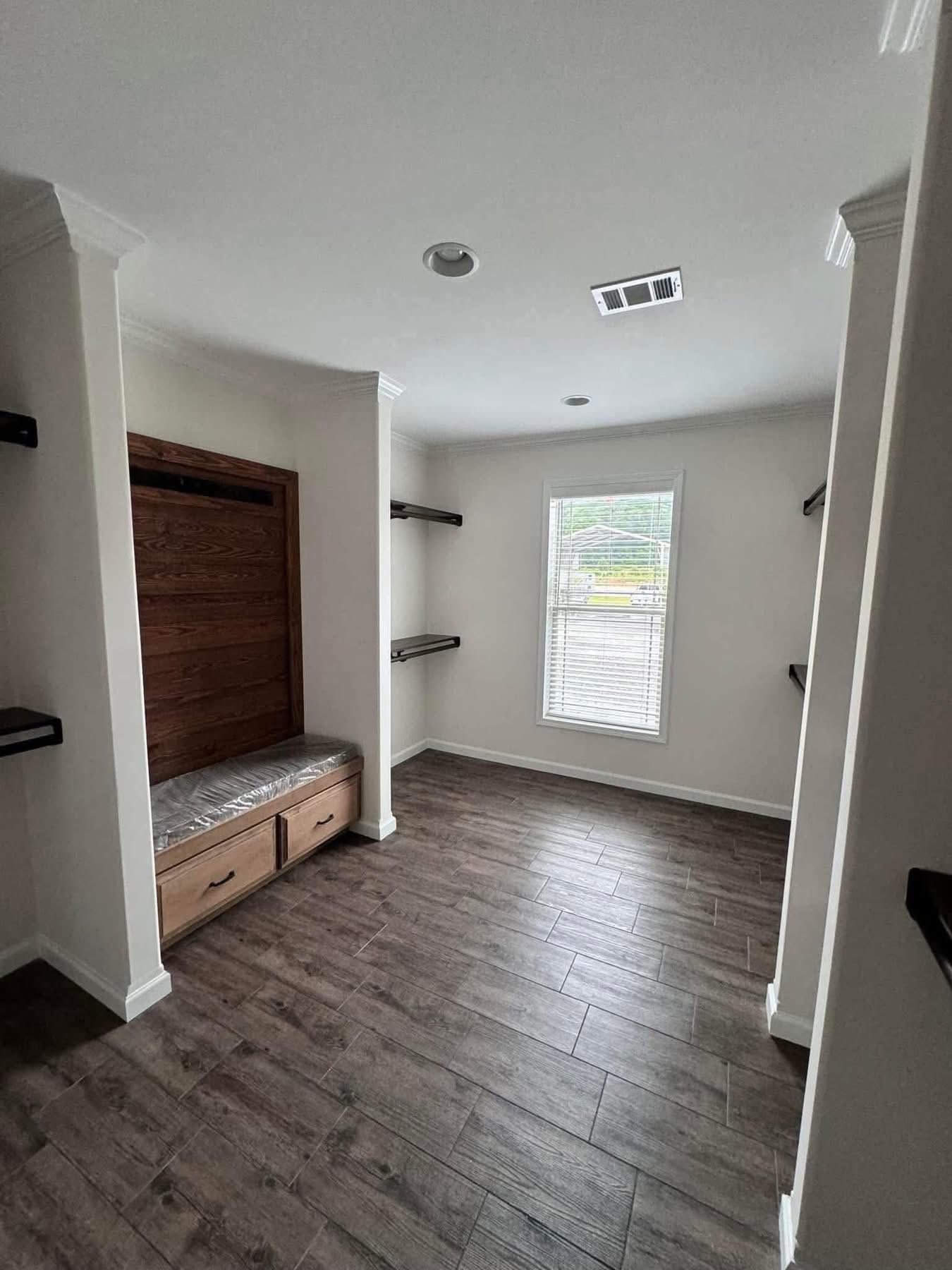 A small room features dark wood flooring, a cushioned bench with drawers, white walls, and black shelves. A window lets in soft daylight, creating a cozy feel.