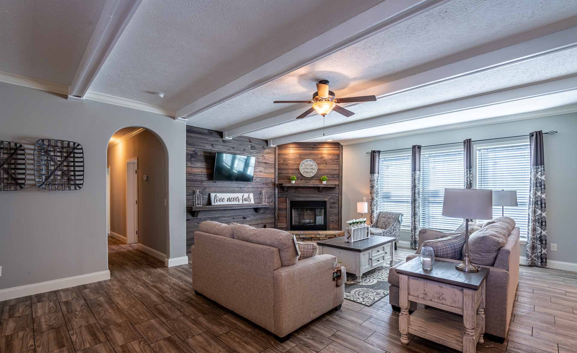 Cozy living room with gray tones, featuring a ceiling fan, beige sofas, rustic wood accents, a mounted TV, and large windows with patterned curtains.