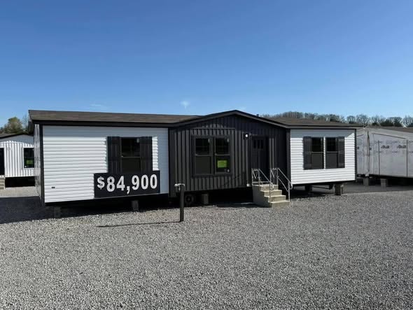 Manufactured home with two-tone siding sits on a gravel lot. A sign on the front displays the price, $84,900. Clear sky and distant trees in the background.