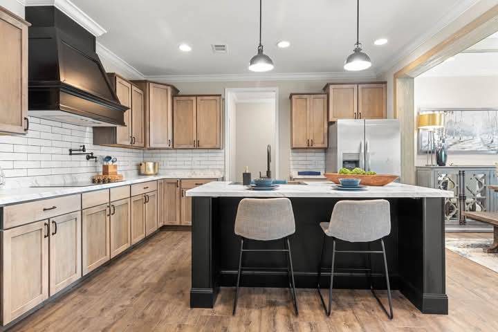 Modern kitchen with light wood cabinets, black range hood, and white subway tile backsplash. A central island has two gray stools, pendant lights above, and a bowl of green apples. The ambiance is warm and inviting.