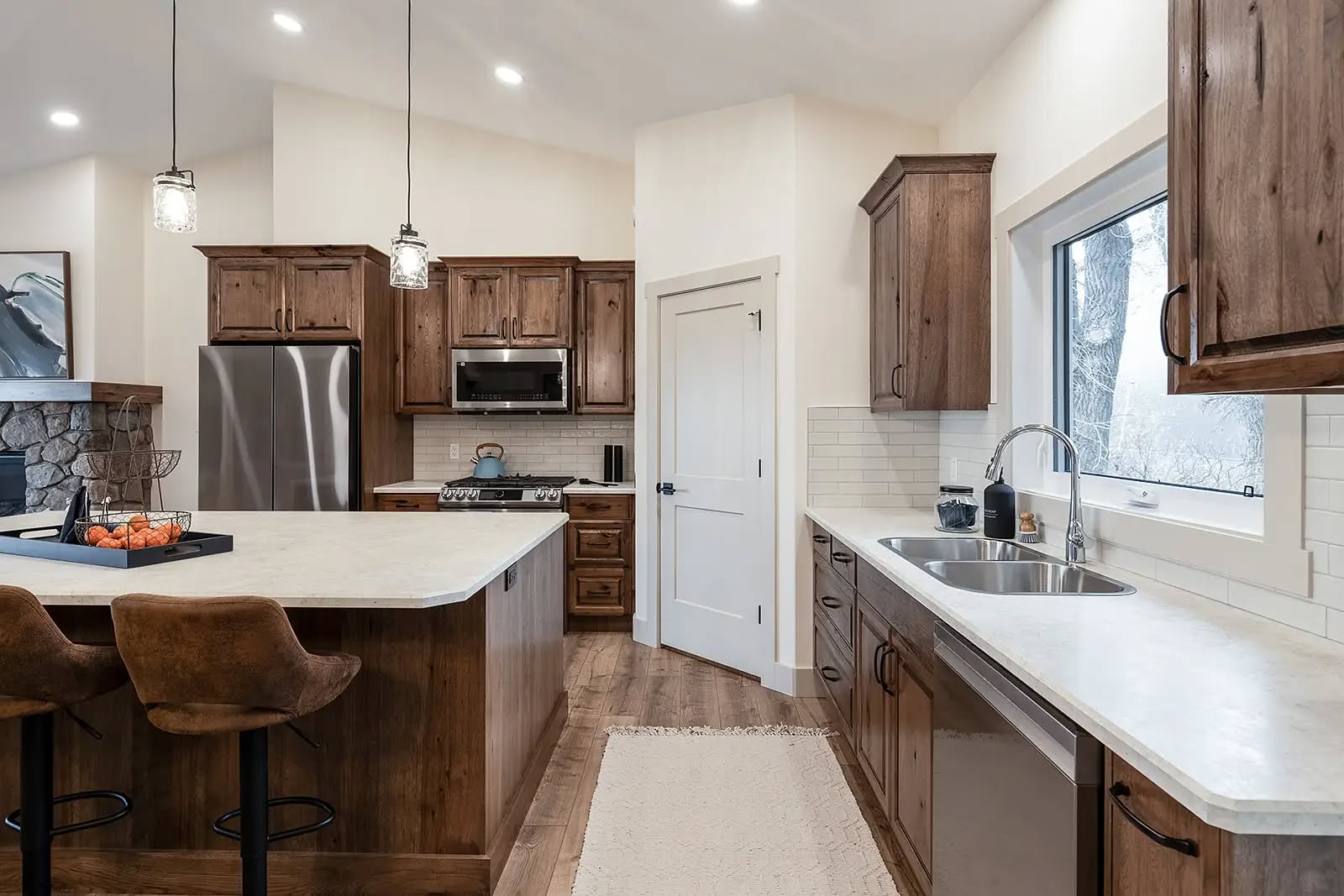 Modern kitchen with wooden cabinets, stainless steel appliances, and a central island with bar stools. Bright lighting and large window create a cozy atmosphere.