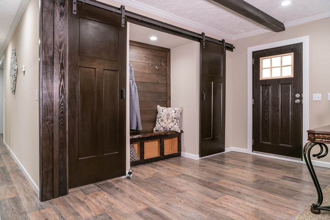 Warm entryway with sliding barn doors revealing a cozy nook with seat, pillows, and coat hook. Dark wood contrasts with light walls and flooring.