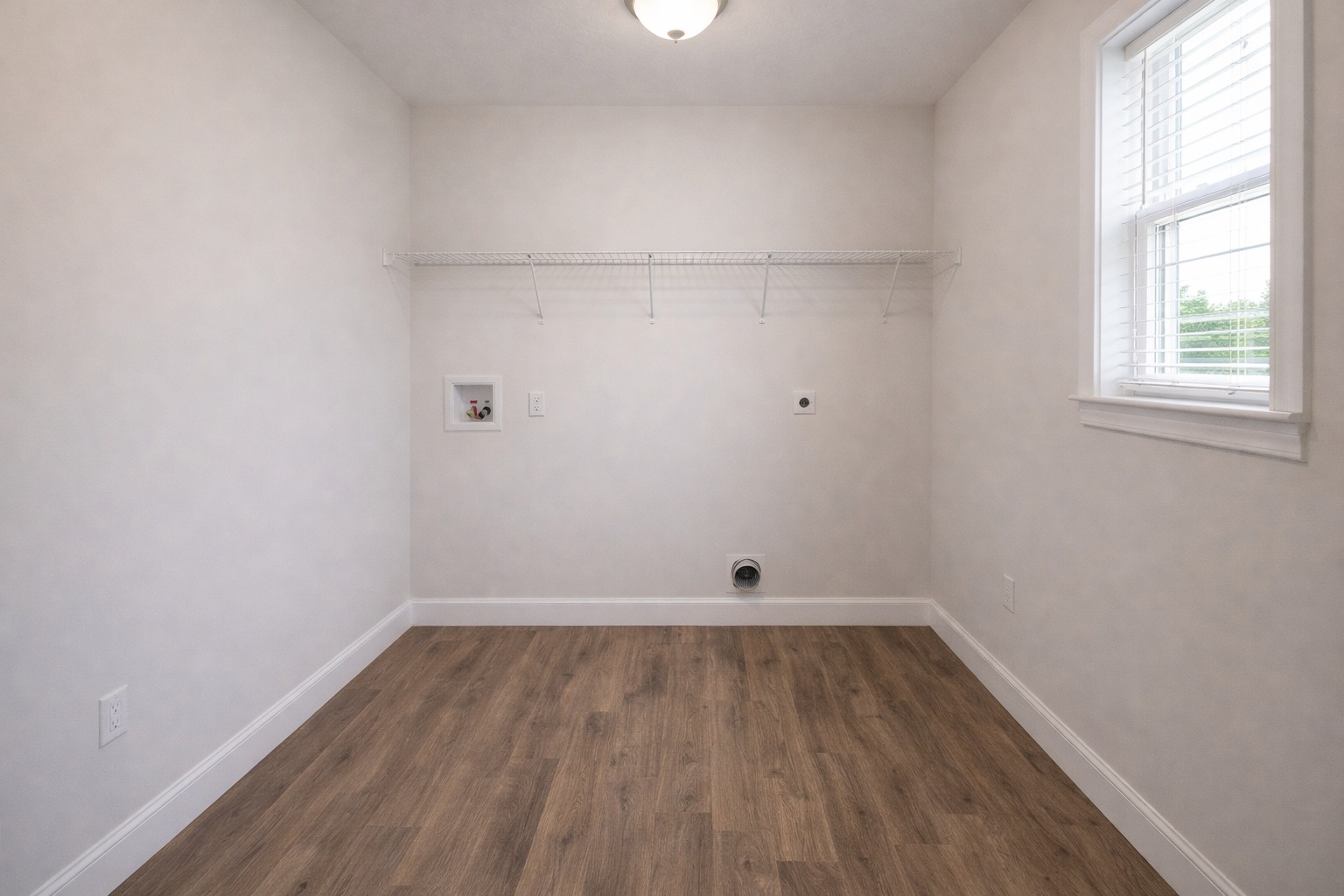 Minimalist laundry room with light beige walls and a wood floor. A wire shelf extends across one wall, under soft overhead lighting. A window to the right brings natural light.