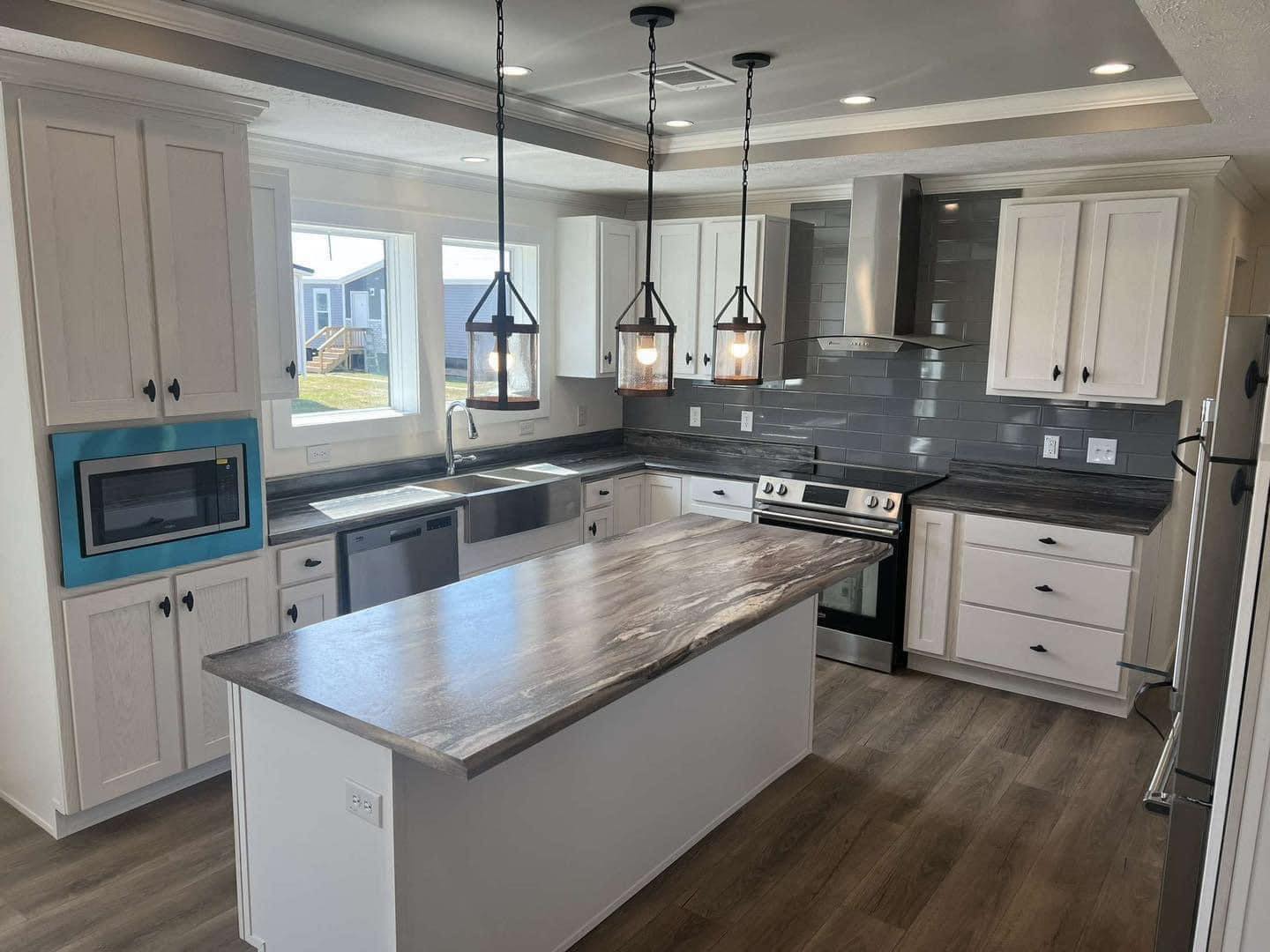 Modern kitchen with gray countertops and white cabinets. Pendant lights hang above a central island. Stainless steel appliances and large windows offer a bright, open atmosphere.
