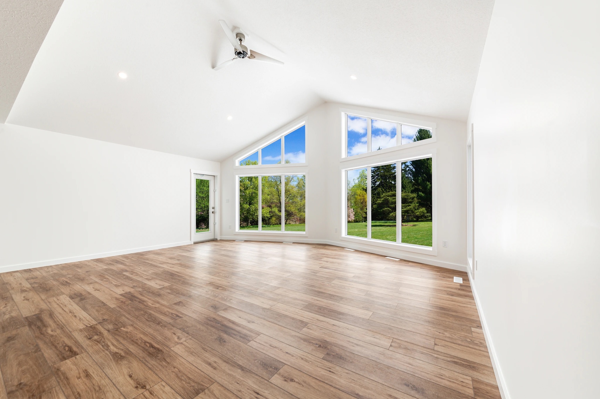 Spacious room with sloped ceiling, wood floors, and large, bright windows overlooking lush greenery and blue sky. Bright, serene ambiance.