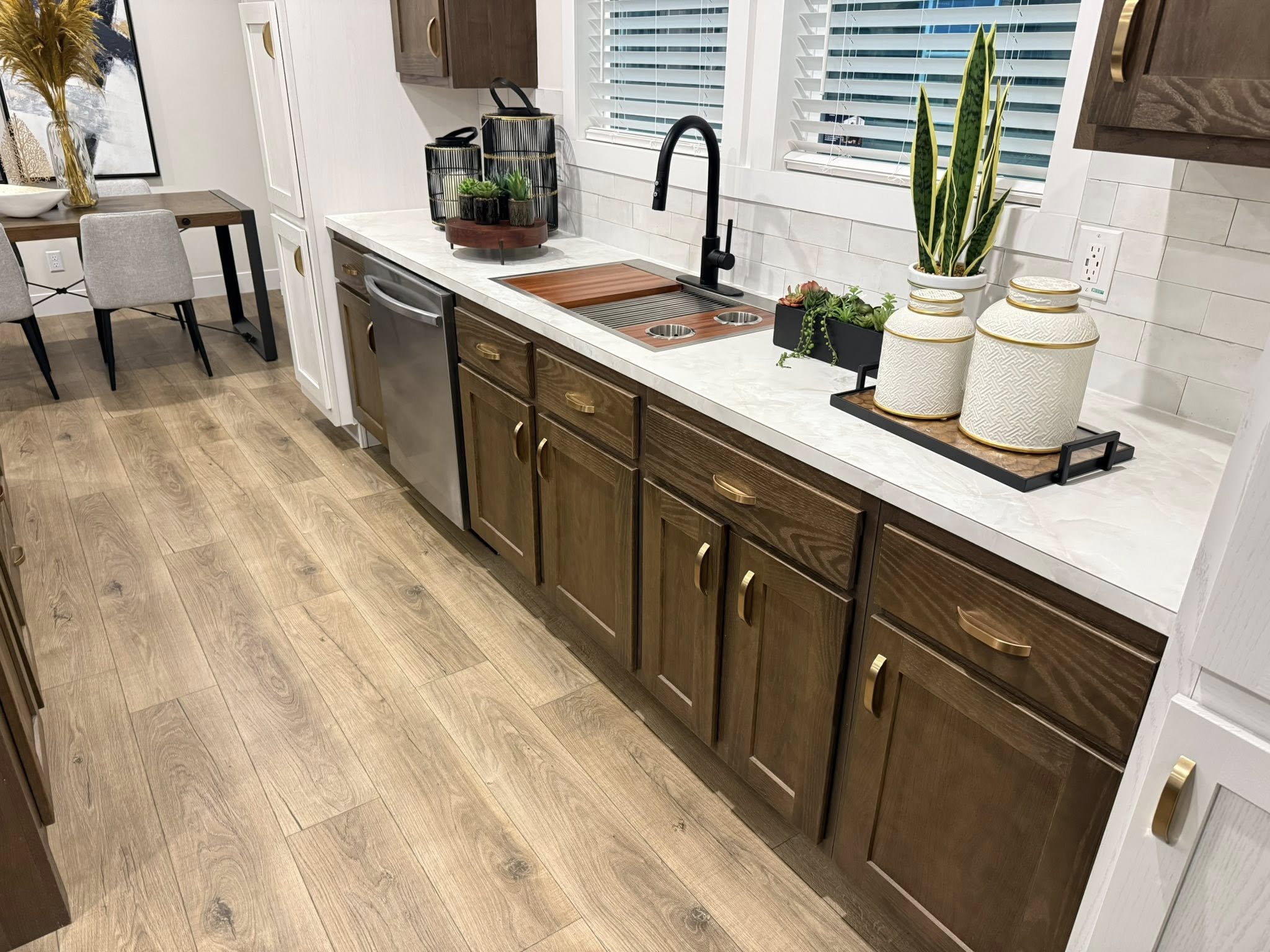 Modern kitchen with wood cabinets, marble countertop, and black faucet. Decor includes potted plants and jars. Dining area visible. Warm, inviting tone.