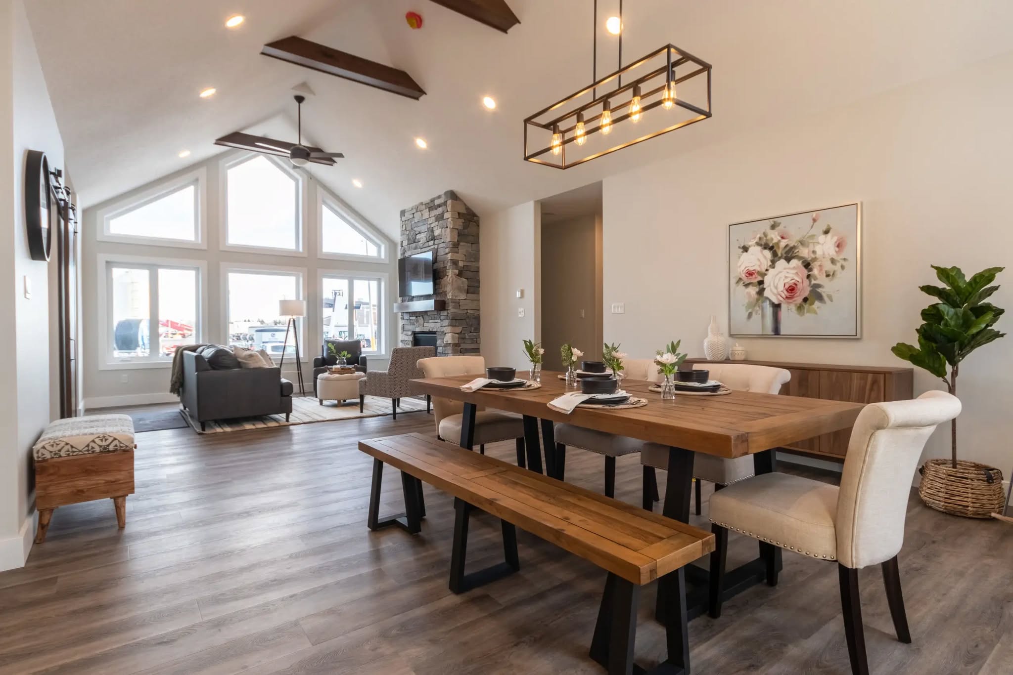 Spacious living area with vaulted ceiling, stone fireplace, and large windows. A wooden dining table, modern light fixture, and floral artwork create a cozy ambiance.