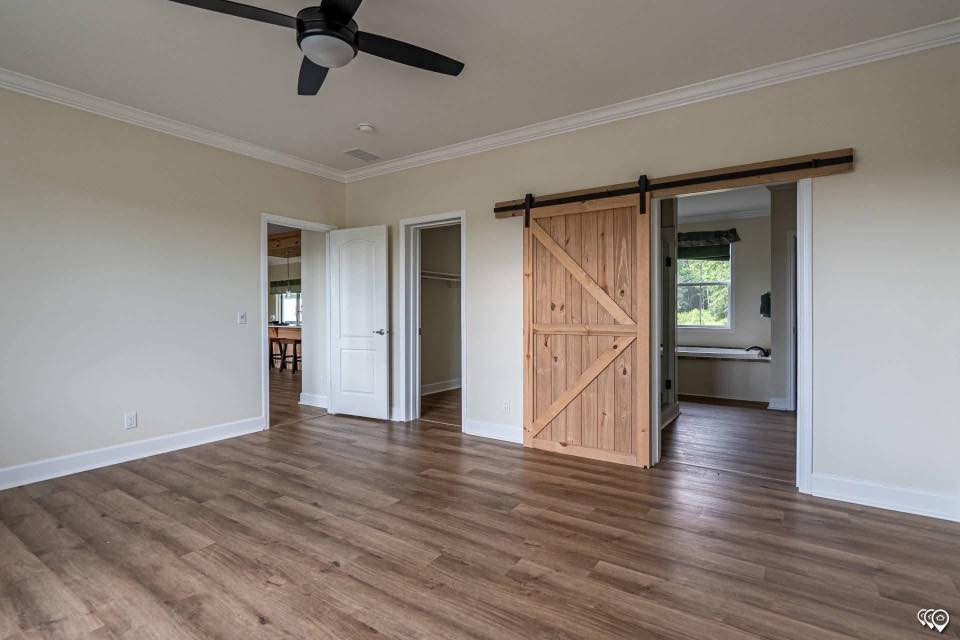 A spacious room with wood flooring, beige walls, and a ceiling fan. It features a sliding wooden barn door and two doorways, creating a warm, rustic feel.