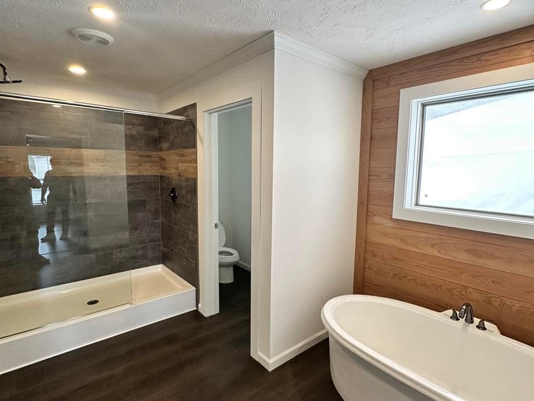 Modern bathroom with dark wood flooring and a stone-tiled shower. A white freestanding tub sits near a wooden wall with a large window, creating a serene feel.
