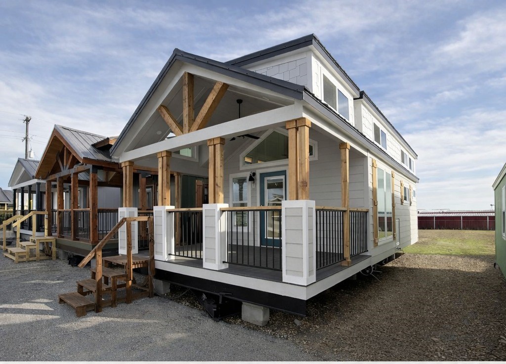 A modern tiny house with a gabled roof and wooden porch supports sits on a gravel lot. It has large windows, a welcoming vibe, and a bright sky above.